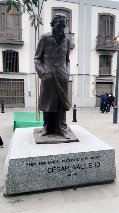 Bronze statue of César Vallejo in a long coat, hands in his pockets, standing on a stone pedestal