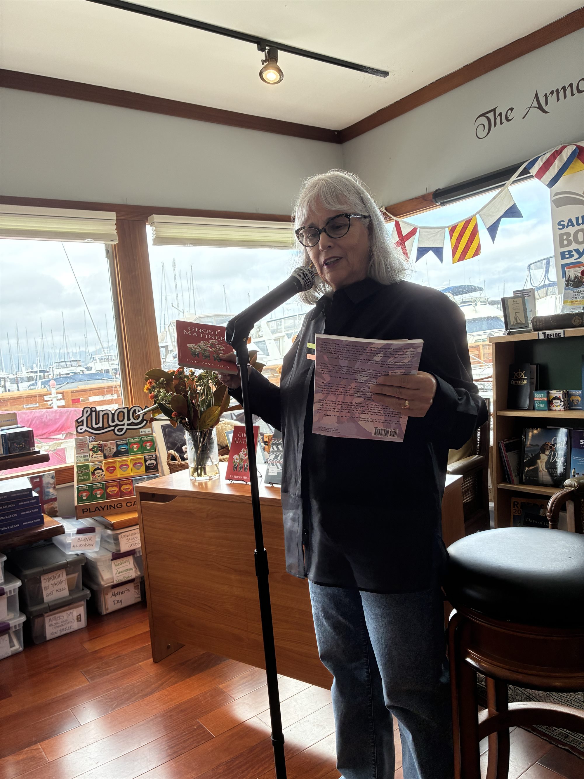 Cathryn Shea reading at a microphone, a copy of Ghost Matinee in her hand, boats visible through the windows