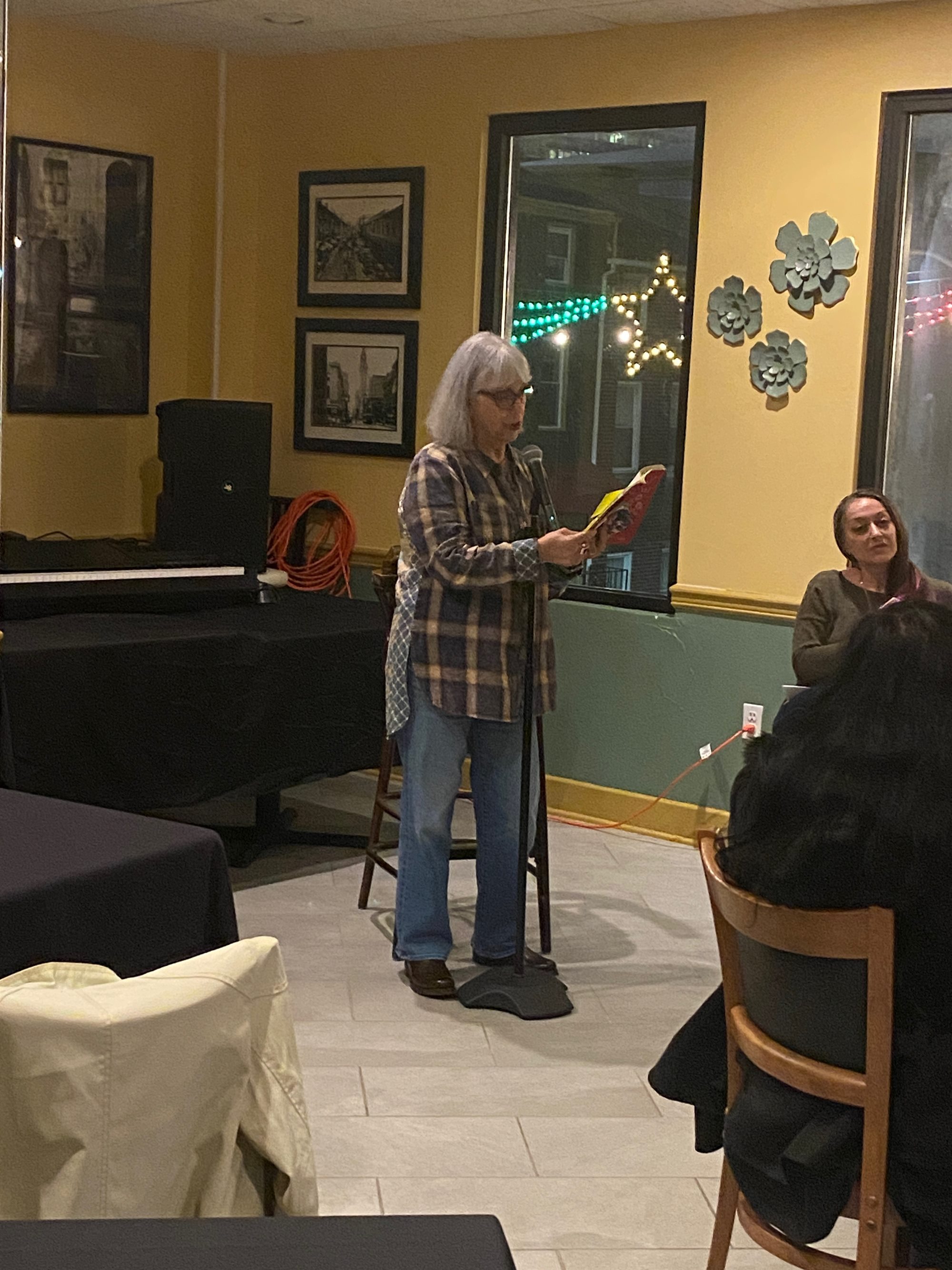 Cathryn Shea reading at a microphone in a yellow-walled room